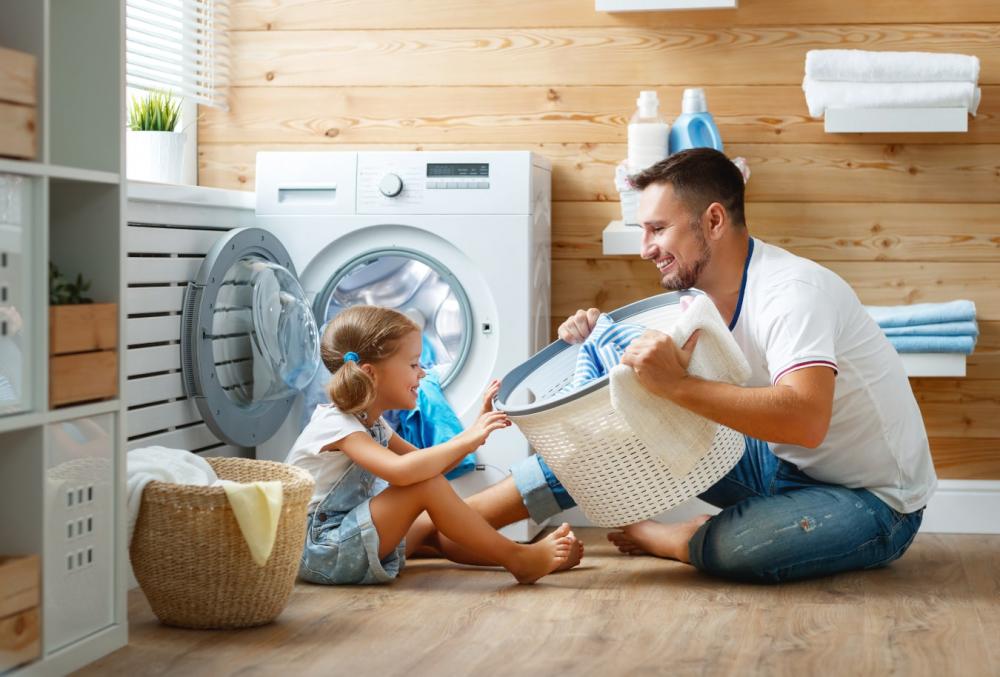 Ein lachender Vater zeigt seiner Tochter den Wäschekorb, gemeinsam haben sie viel Spaß an der Waschmaschine