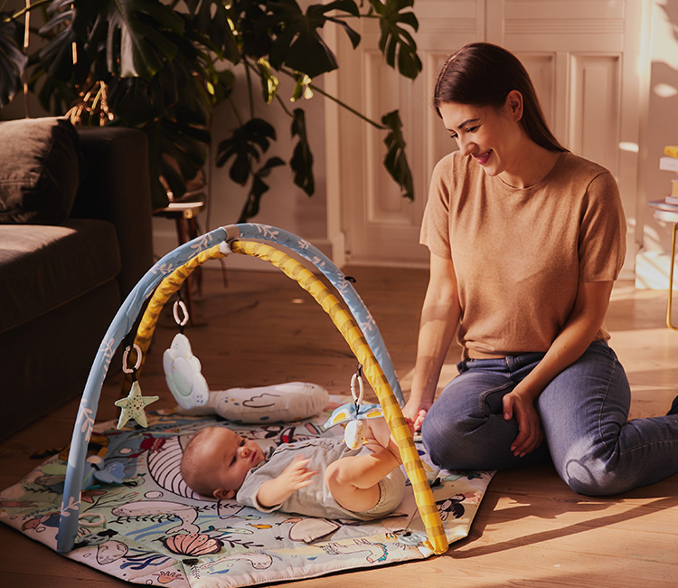 Das Baby liegt auf dem Rücken auf der Spielmatte von Kinderkraft im Wohnzimmer. Neben ihm sitzt die Mutter und lächelt das Baby an.