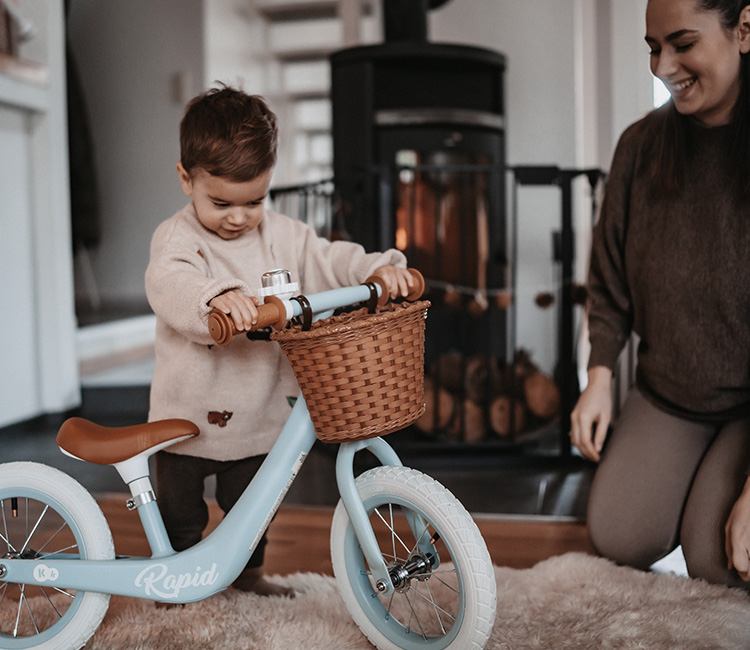 Der 2-jährige Junge spielt im Wohnzimmer mit einem Laufrad, begleitet von seiner Mutter, die in der Nähe ist.