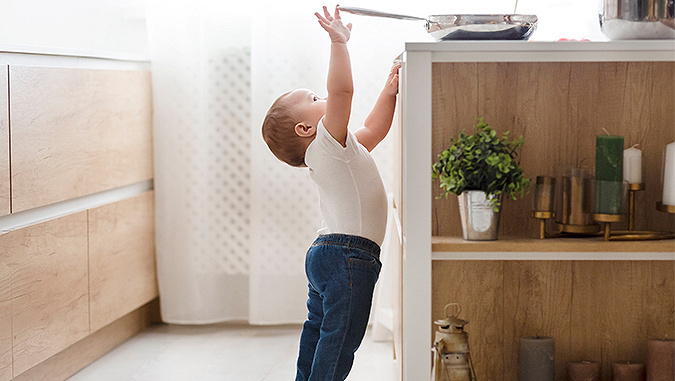 Ein kleines Kind, bekleidet mit Jeans und weißer Bluse, steht in der Küche. Sie stellt sich auf die Zehenspitzen und greift mit der Hand nach einer auf dem Tresen stehenden Pfanne.