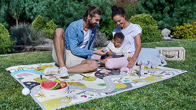 Auf der Wiese an einem sonnigen Tag sitzen die lächelnden Eltern und spielen mit ihrem kleinen Kind. Daneben liegen Spielzeug, ein Teller mit Obst und ein Picknickkorb.