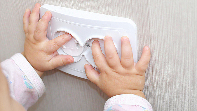 Die Hände des Kindes ruhen auf einer elektrischen Steckdose. Sie versucht, ihre Finger hineinzustecken, aber die Steckdose hat Sicherheitsvorrichtungen, die dies unmöglich machen.