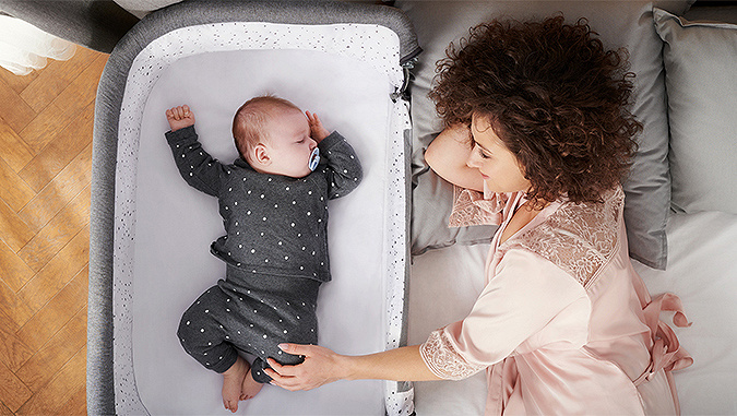 Im Schlafzimmer blickt die im Bett liegende Mutter lächelnd auf und hält das Bein des winzigen Babys, das im daneben stehenden Kinderkraft-Bettchen schläft.