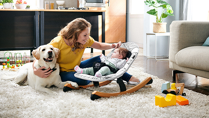 In der Wohnung liegt ein kleines Kind auf einem Kinderkraft-Schaukelstuhl. Daneben streichelt die Mutter mit einer Hand den Kopf des Kindes und umarmt mit der anderen einen großen Hund.