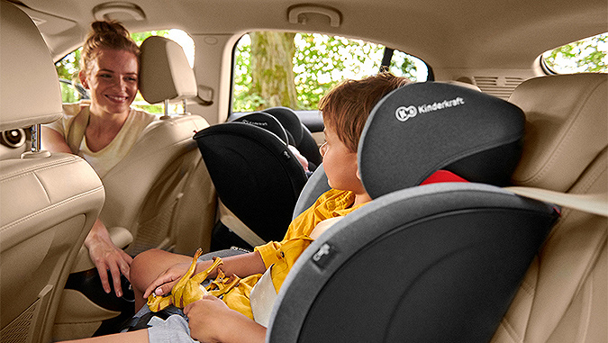 Im Auto eine Mutter und zwei Kinder in Kinderkraft-Autositzen. Das Neugeborene schläft rückwärts, während die Mutter sich dem kleinen Jungen zuwendet.