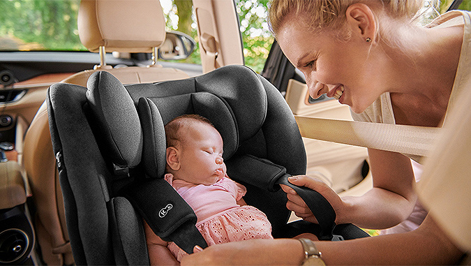 Ein neugeborenes Baby schläft in dem grauen Kinderkraft-Sitz, der verkehrt herum im Auto platziert ist, während seine lächelnde Mutter den Sicherheitsgurt anlegt.