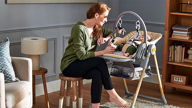 Die lächelnde Mutter sitzt auf einem kleinen Stuhl im Zimmer. Sie hält den Fuß ihres Babys, das neben ihr in einem Kinderkraft-Stuhl schläft.