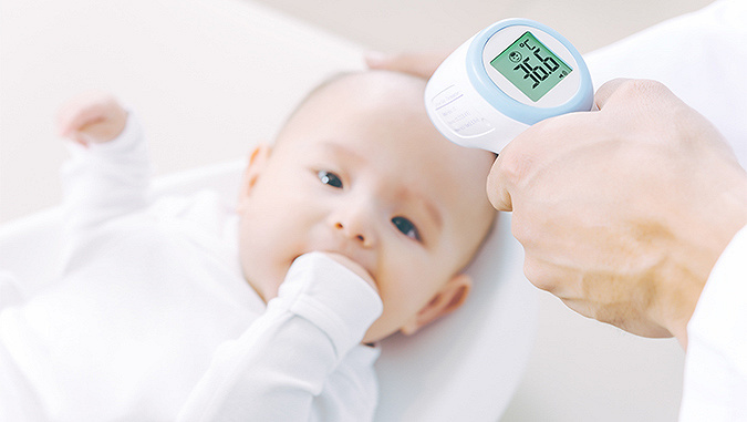 Ein Baby liegt beim Arzt und hält die Hand in den Mund. Der Arzt misst seine Temperatur mit einem berührungslosen Thermometer - sie beträgt 36,6°C.
