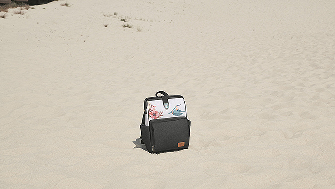 An einem sonnigen Tag am Sandstrand liegt ein weiß-grauer Kinderkraft-Rucksack mit einem Blumen- und Vogelmotiv.