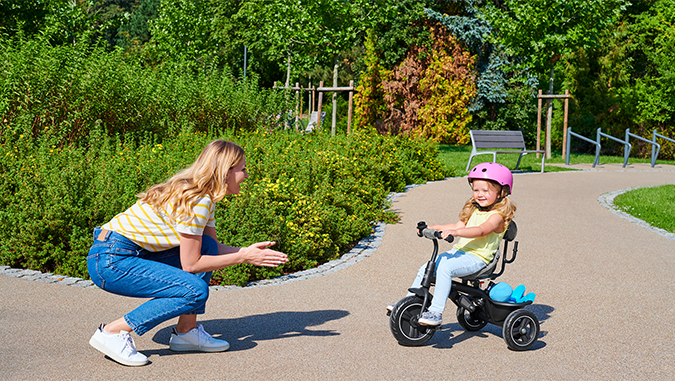 Auf einem Weg im Park fährt an einem sonnigen Tag ein Mädchen mit einem rosa Helm auf einem Kinderkraft-Dreirad in Richtung seiner lächelnden Mutter.
