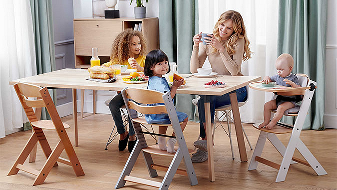 Die Mutter und drei Kinder frühstücken am Tisch sitzend. Zwei kleinere Kinder sitzen in speziellen Kinderkraft-Stühlen.