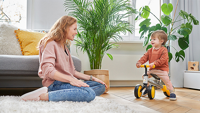 Ein kleiner Junge fährt auf einem Kinderkraft-Dreirad durch den Raum. Er wird von seiner lächelnden Mutter beobachtet, die neben ihm auf dem Boden sitzt.