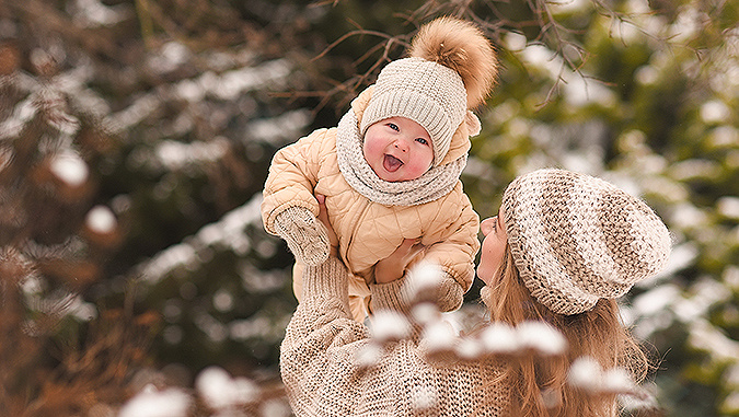 Winter, Sie können Schnee auf den Bäumen im Hintergrund sehen. Mama in Wollpullover und Mütze nimmt ein lachendes Kind in Mütze und Winteranzug auf den Arm.