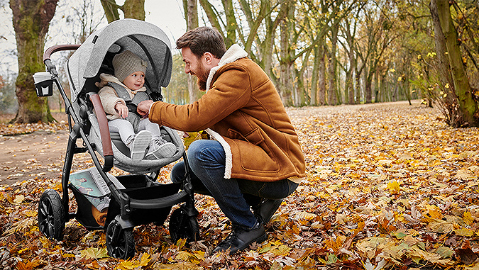 In einem Park voller Herbstlaub beugt sich ein lachender Vater über den Kinderkraft-Kinderwagen und passt den Sicherheitsgurt des darin sitzenden Mädchens an.