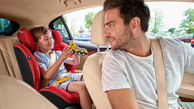 Im Auto wendet sich der Vater - der Fahrer - einem wenige Jahre alten Jungen zu, der hinter ihm auf dem Sitz der Marke Kinderkraft sitzt und spielt.