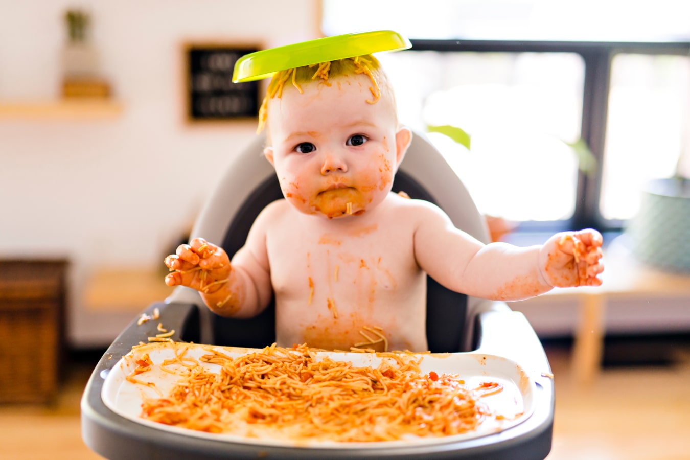 Ein Baby, schmutzig von Spaghetti, sitzt auf einem Fütterungsstuhl, hat einen Teller mit Essen auf dem Kopf
