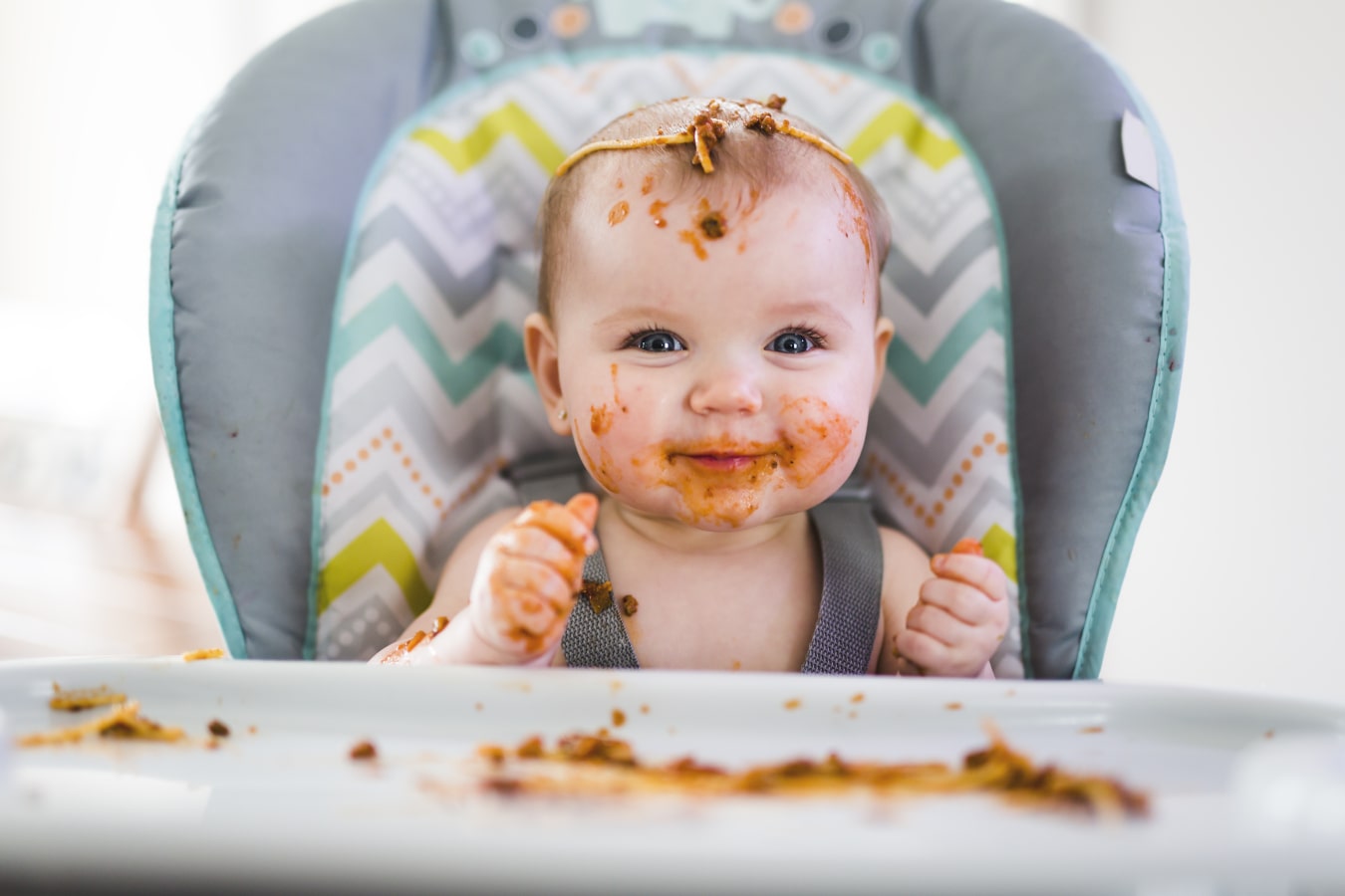 Ein Baby, schmutzig von Spaghetti, sitzt angeschnallt in einem Essensstuhl. Er ist glücklich und lächelt, er hat reichlich Futter dabei und auf dem Tablett.