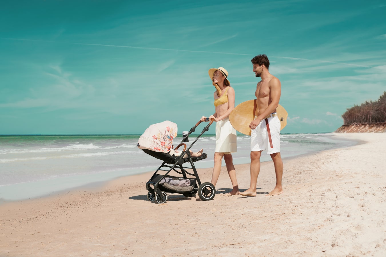 Mutter und Vater gehen mit einem Kind im schwarzen Buggy mit einem beigen Verdeck am Strand spazieren 
