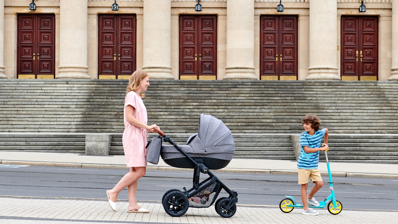Eine Mutter geht mit einem klassischen, grauen Kinderwagen und einem Kind auf einem zweirädrigen, türkisfarbenen Tretroller in der Stadt spazieren
