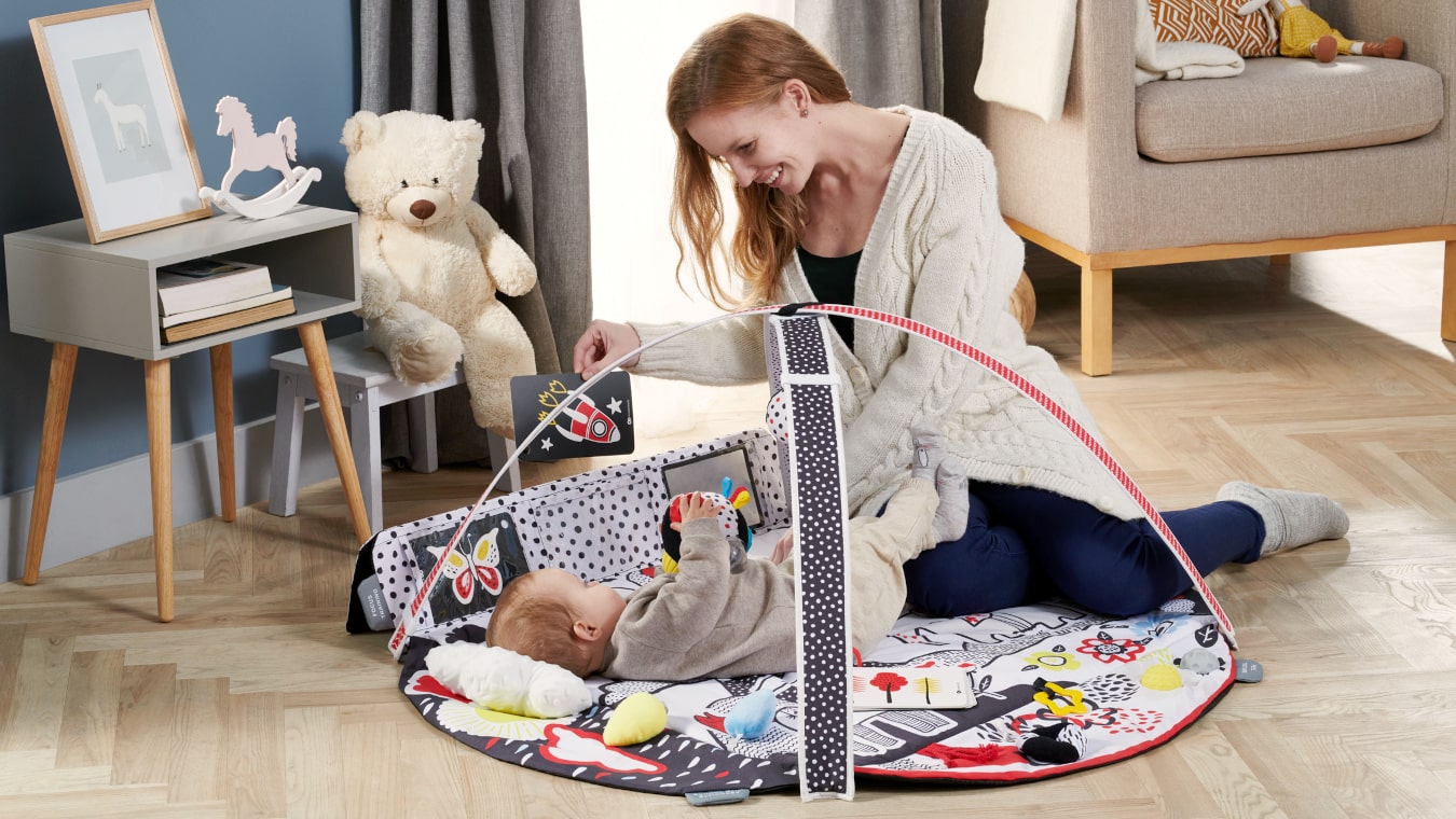 Eine lächelnde Mutter spielt in der Wohnung mit dem Kind, das auf einer bunten sensorischen Matte liegt, und zeigt ihm ein kontrastreiches Bild