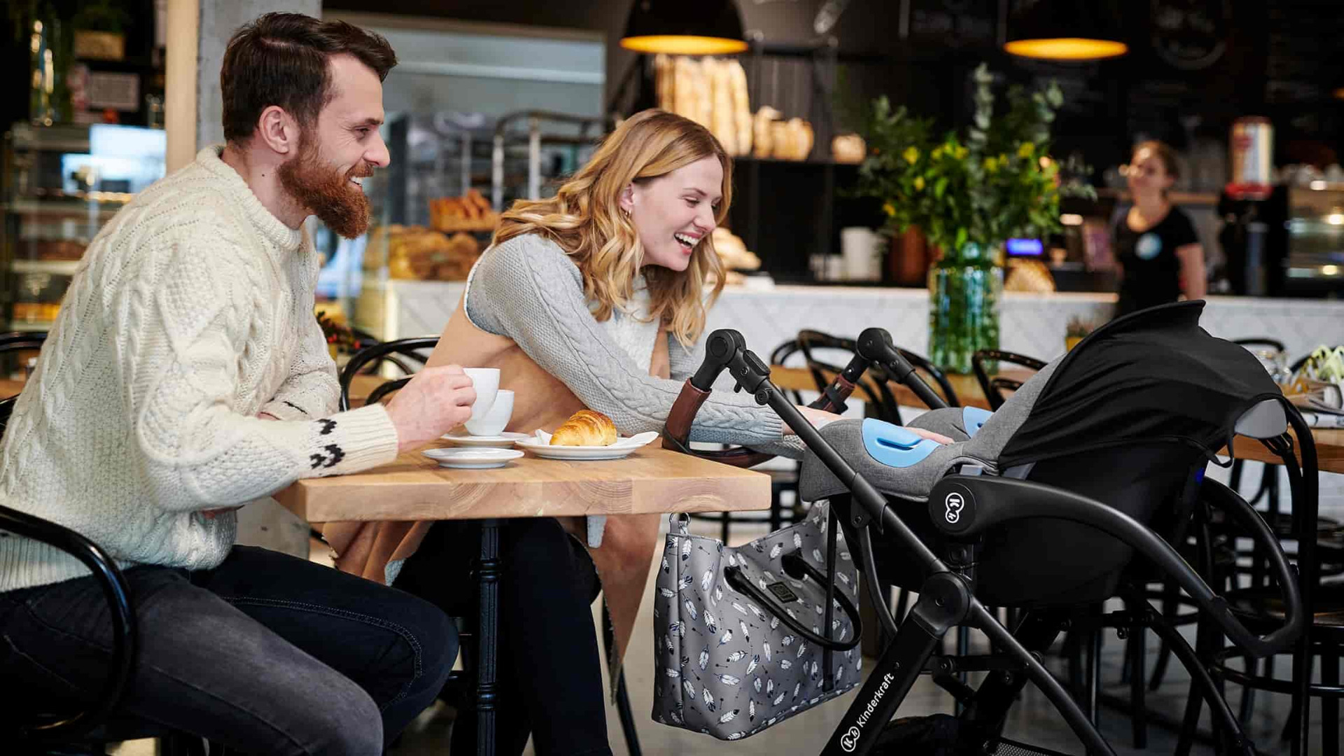 Ein lachendes Paar sitzt in einem Café. Die Frau greift in den XMOOV-Wagen. Der Mann hält eine Tasse Kaffee in der Hand.