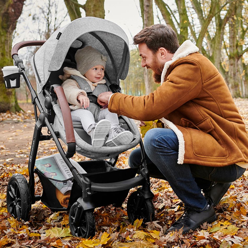 Ein Vater beugt sich in einer Jacke zum Kind, das im Kinderwagen sitzt. Das Kind lacht, um sie herum liegen Herbstblätter.