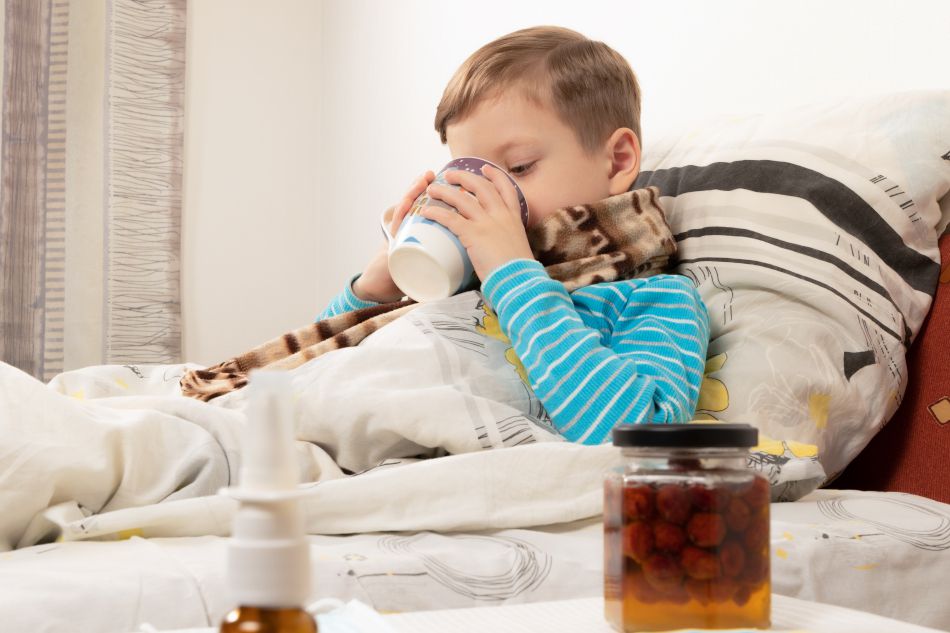 Ein kranker Junge trinkt aus der Tasse warmen Tee. Auf dem Tisch steht ein Glas mit Himbeersirup gegen Erkältungen