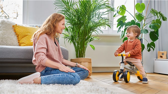 Die Mutter lächelt das Kind an, das mit dem Dreirad Cutie Kinderkraft fährt. Sie sind zu Hause, im Hintergrund sind grüne Pflanzen und ein graues Sofa zu sehen. Das Kind lächelt.