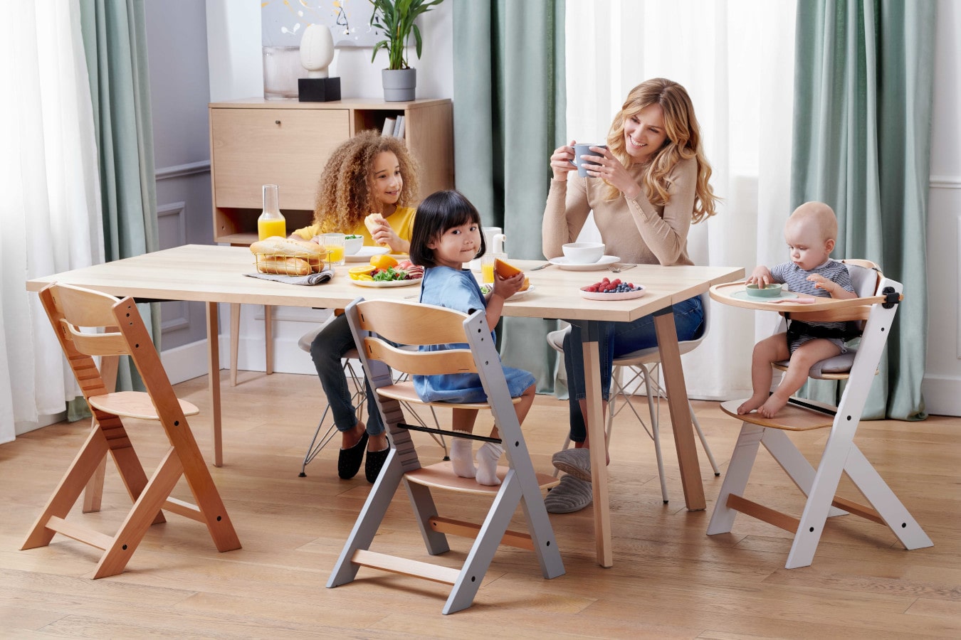 Mutter und drei Kinder sitzen am Tisch, die Mutter trinkt Kaffee, die Kinder freuen sich. Der ENOCK-Kinderhochstuhl ist in 3 Konfigurationen erhältlich - vom Kinderhochstuhl bis zum Stuhl für ältere Kinder.