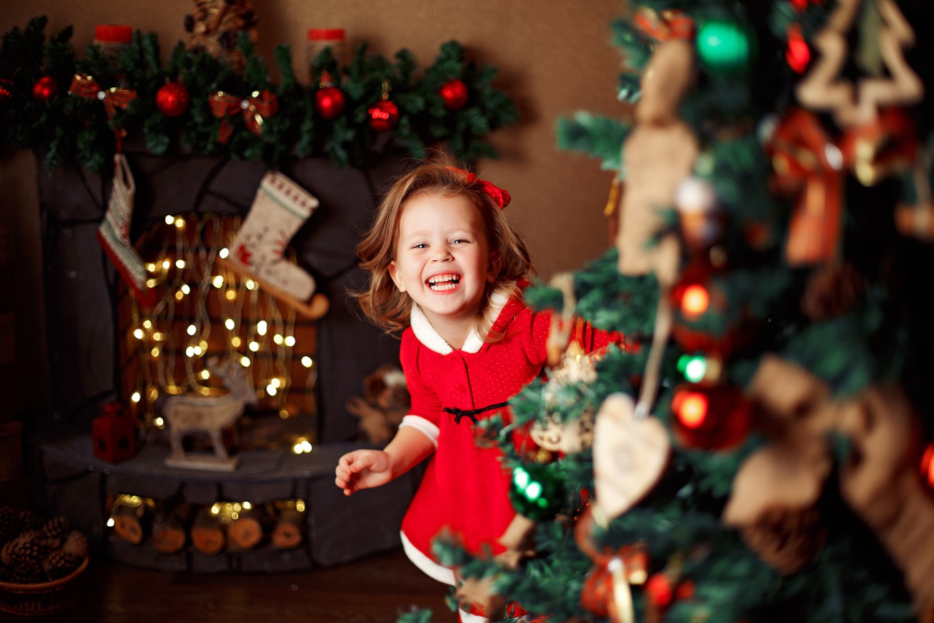 Ein fröhliches Mädchen im roten Kleid blickt hinter einem Weihnachtsbaum hervor, es  lacht dabei und ist glücklich.